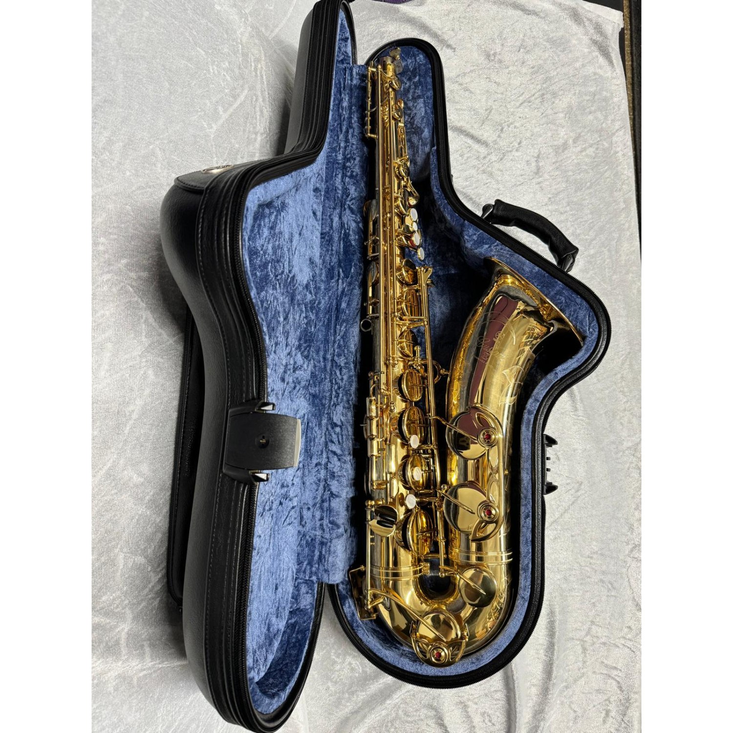 Tenor saxophone case laying open with saxophone inside, showing blue velvet lining; against white velvet background